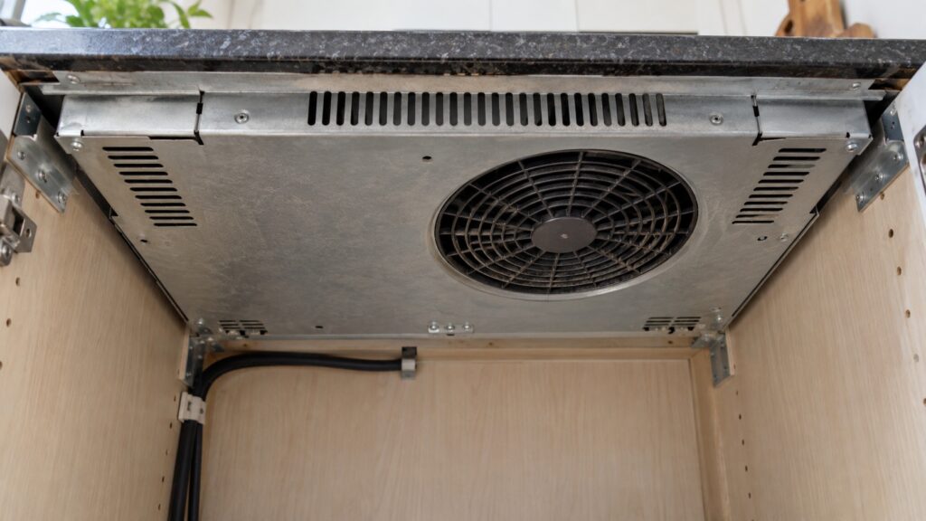 The underside of an induction cooktop seen from inside the cabinet below showing the cooling fan intake