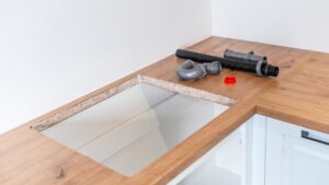 A rectangular cutout in a wooden kitchen countertop with the raw chipboard edge exposed and fittings lying beside it